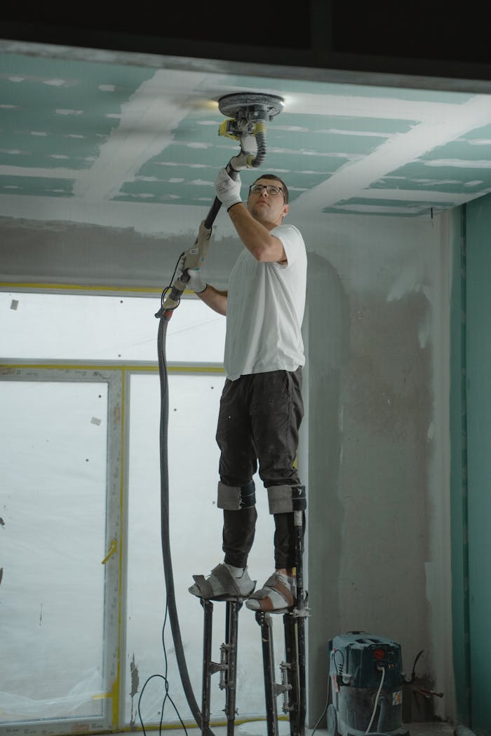 A man on stilts uses a ceiling sander for home renovation, focusing on drywall preparation.